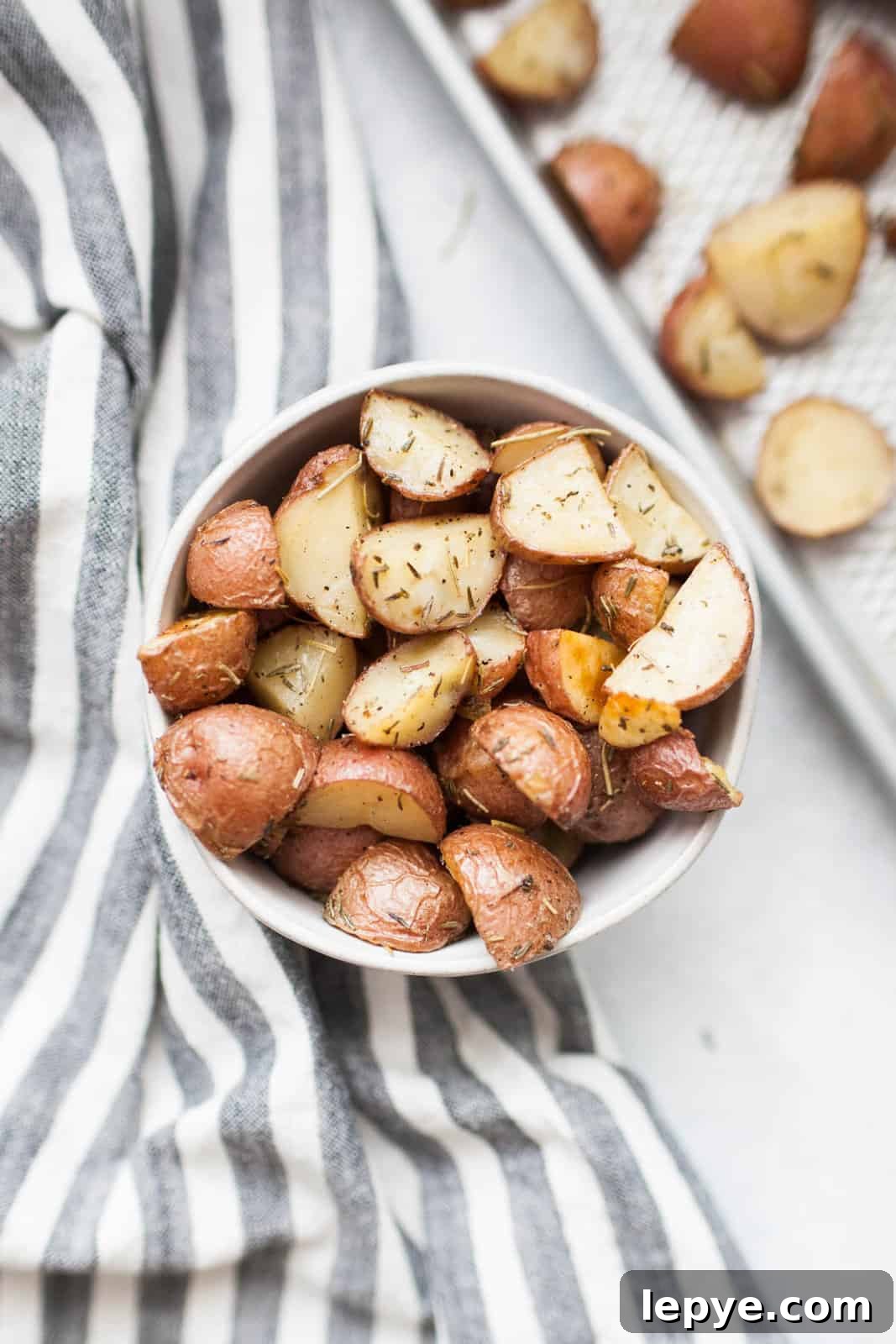A bowl of rosemary potatoes seasoned with herbs sits on a striped cloth, with a tray of more roasted potatoes in the background.