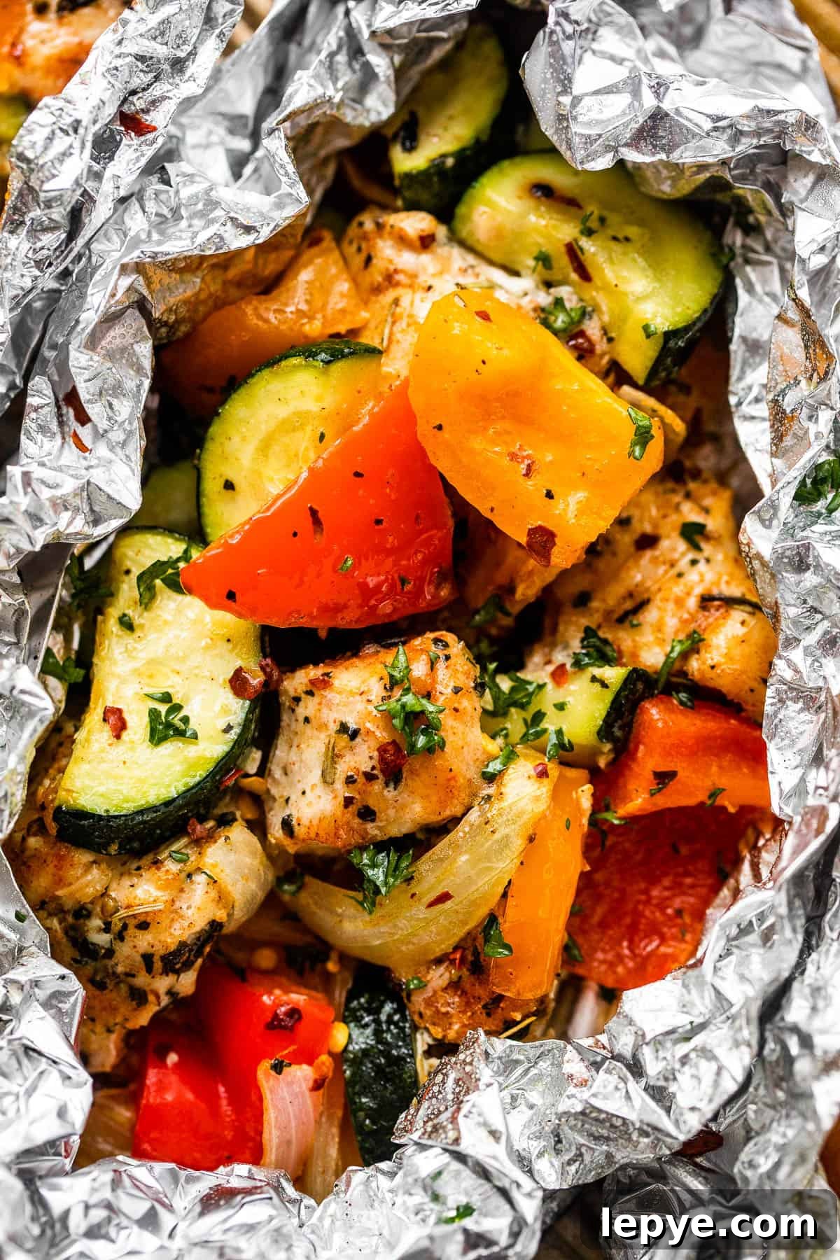 Chicken and Vegetables in Foil Packets combine juicy chicken and crisp veggies, all cooked together in foils for a convenient family meal. up close shot of diced chicken, sliced zucchini, and sliced peppers all layered in an aluminum foil packet.