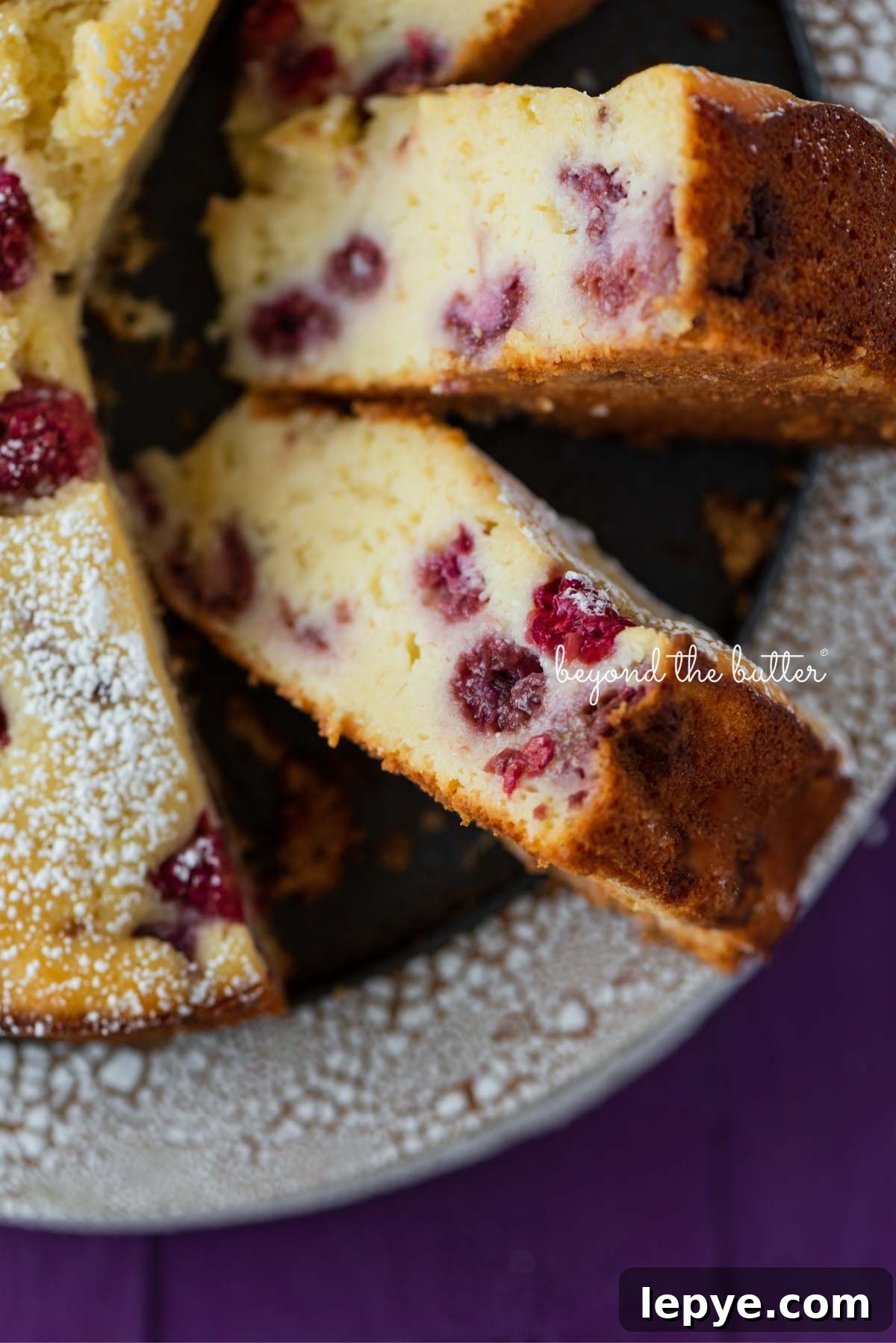 Lemon raspberry ricotta cake dusted with powdered sugar on crackled ceramic cake stand and purple wood background.