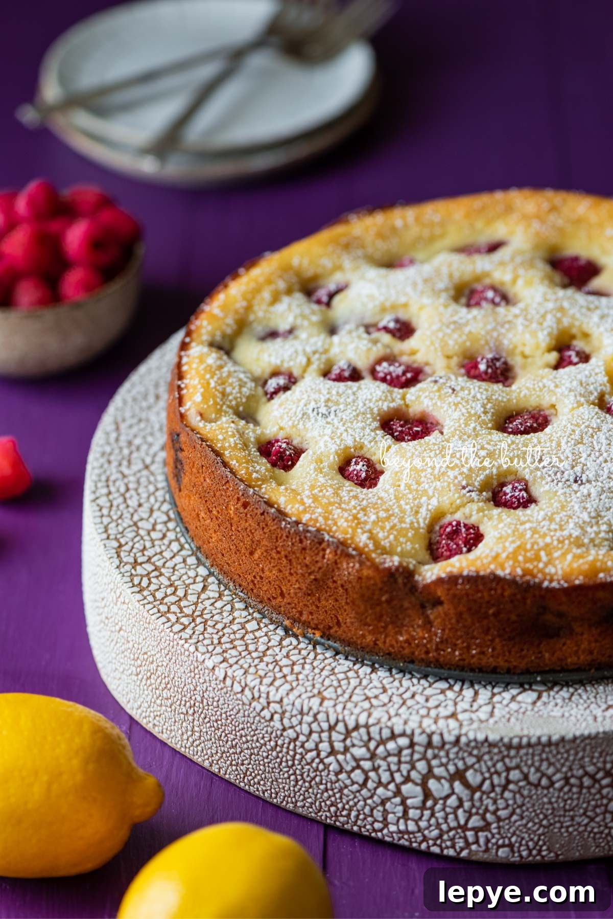 Lemon raspberry ricotta cake dusted with powdered sugar on crackled ceramic cake stand and purple wood background.