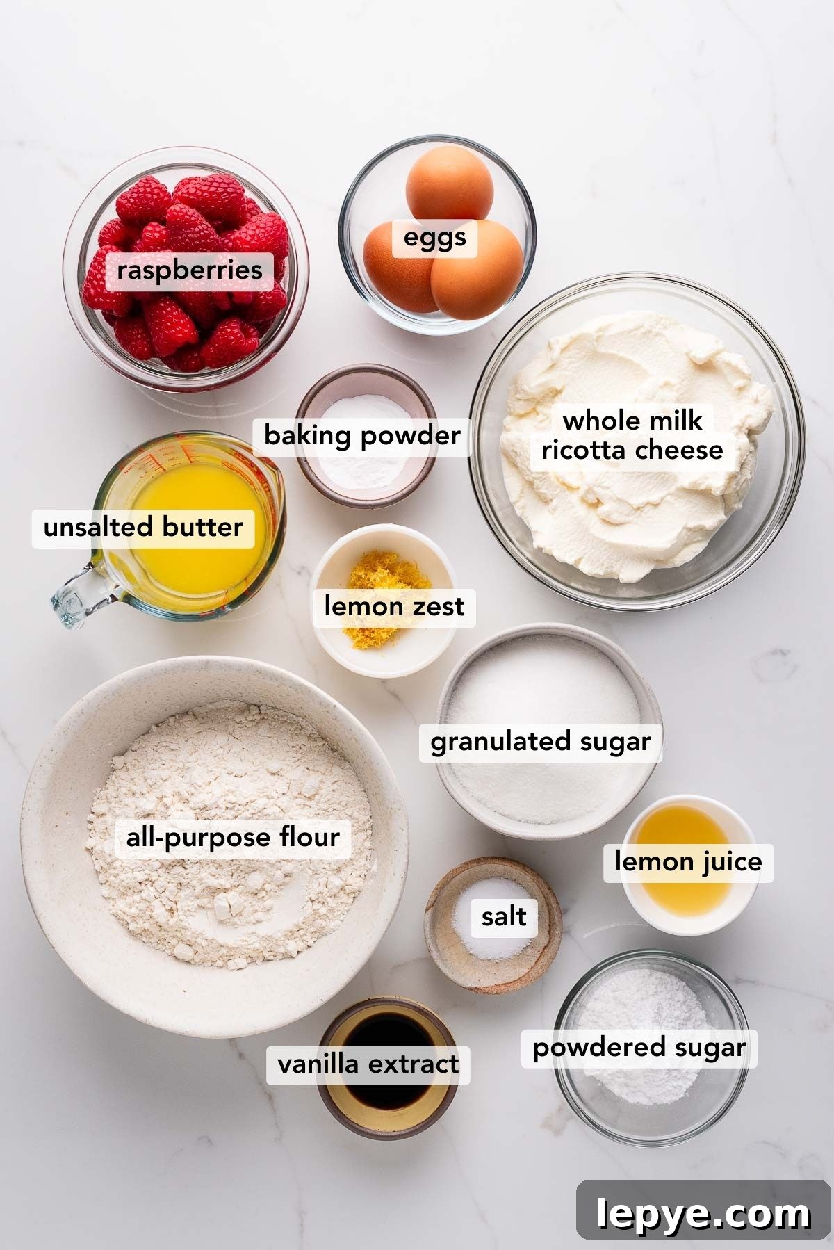 Labeled ingredients in bowls used to make lemon raspberry ricotta cake on a white marbled background.