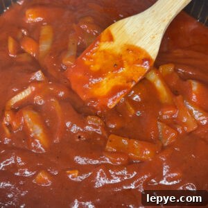 A close-up of a pan filled with tomato sauce, BBQ sauce, and sliced onions, being stirred with a wooden spatula.