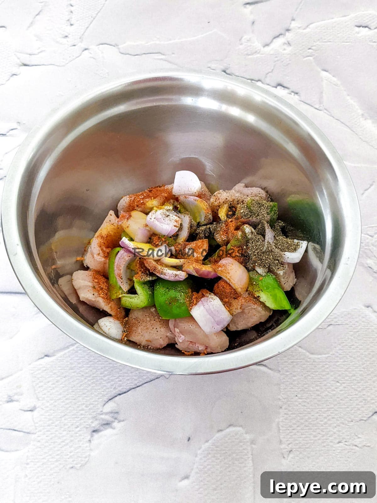 Spices and oil on top of peppers, onion and chicken cubes in a steel bowl.