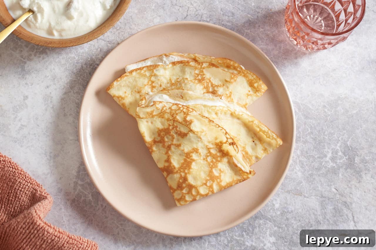 Sweet Nalysnyky: Classic Ukrainian Crepes with Cottage Cheese Filling 10 Ukrainian crepes on a pink plate, a bowl of ricotta cheese filling is in the upper left, a pink glass is in the upper right.