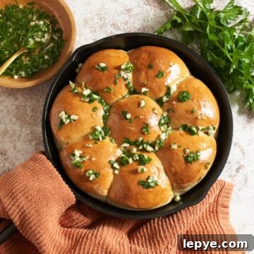 Pampushky (Ukranian garlic bread) in a cast iron skillet topped with a garlic-herb oil. A bowl of the oil is to the left, fresh parsley is on the right, the skillet handle is wrapped in an orange towel.