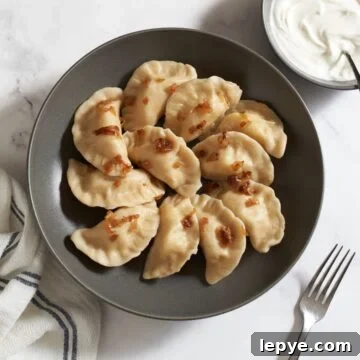 A bowl of Ukrainian potato varenyky dumplings topped with caramelized onions. A bowl of sour cream and a fork are on the right, a blue and white striped towel is on the left.