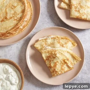 Sweet Nalysnyky: Classic Ukrainian Crepes with Cottage Cheese Filling 14 Two Ukrainian crepes with a sweet cheese filling folded on a pink plate, two more plates of stacked and folded crepes are above it, a bowl of filling is on the left.