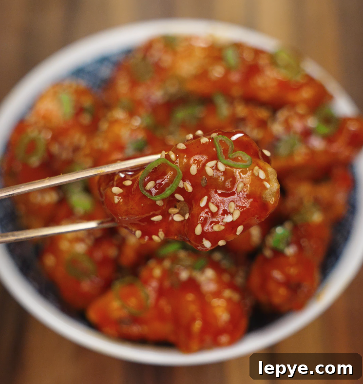 A single piece of spicy Korean fried chicken garnished with sesame seeds and scallions being held with chopsticks.