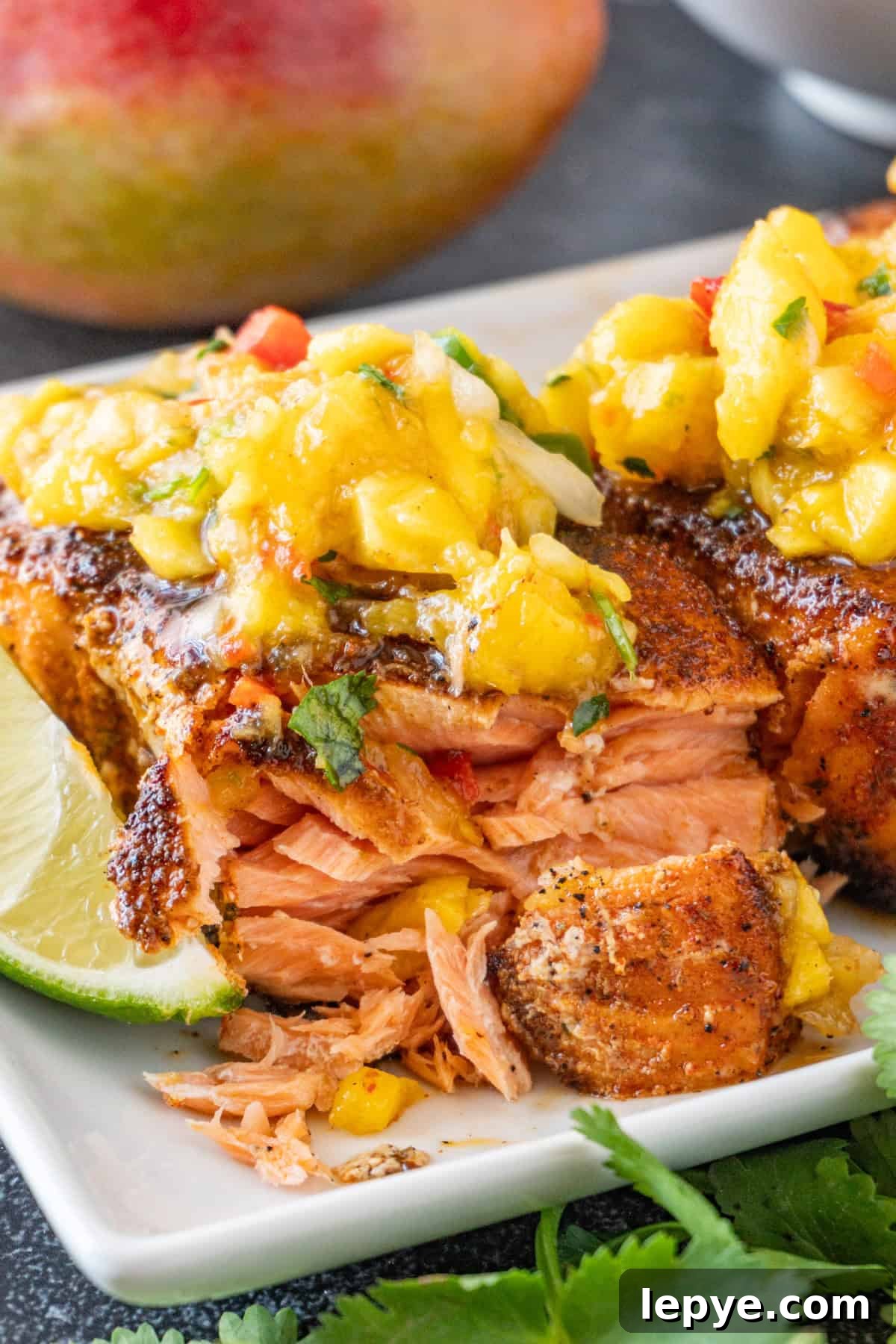 Grilled Salmon with Fresh Mango Salsa Recipe 3 Salmon cut open to show cooked flaky layers, topped with mango salsa, sitting on a white plate.