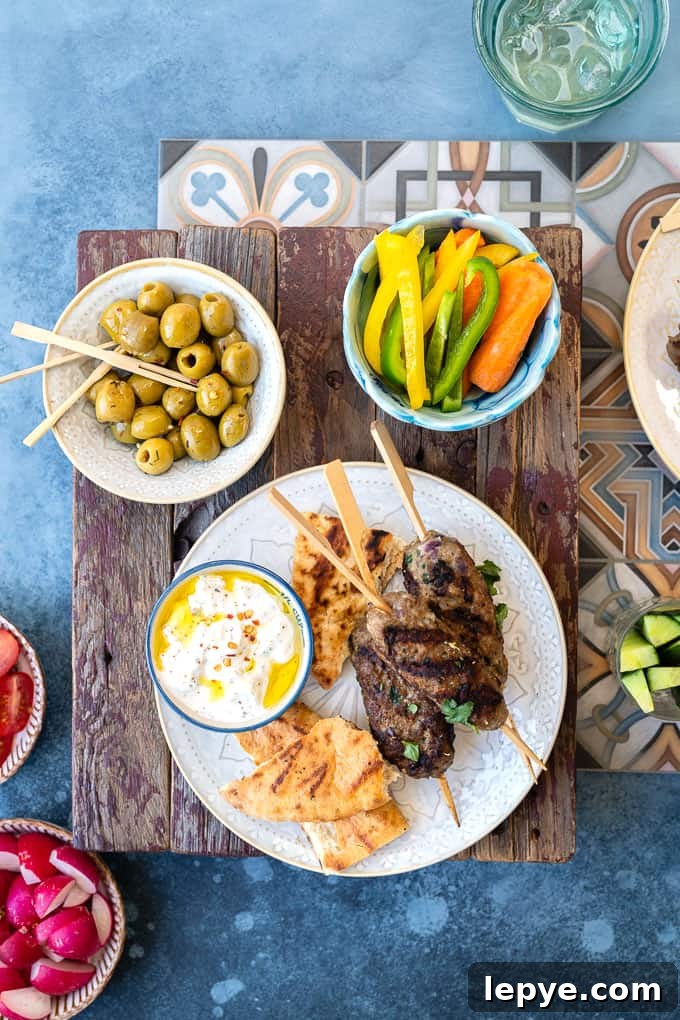 Authentic Lamb Kofta Recipe: Juicy Middle Eastern Meatballs 3 Lamb kofta on a plate with pitta bread and tzatziki surrounded by small plates of vegetables and olives