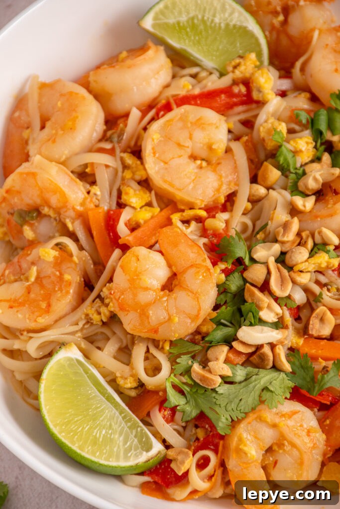 Close up of shrimp pad thai in a bowl with rice noodles, veggies, and garnished with cilantro, lime wedges, and chopped peanuts.