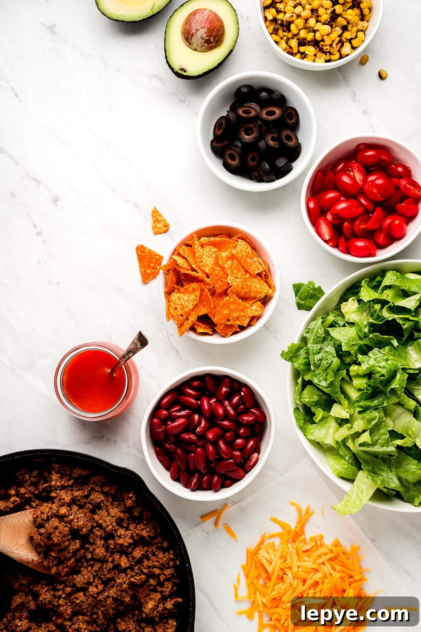 Dorito Taco Salad Recipe: Crunchy Tex-Mex Dinner Idea 5 Ingredients on a marble surface- roasted corn, avocado, black olives, tomatoes, Doritos, lettuce, kidney beans, shredded cheese, taco meat, and dressing.