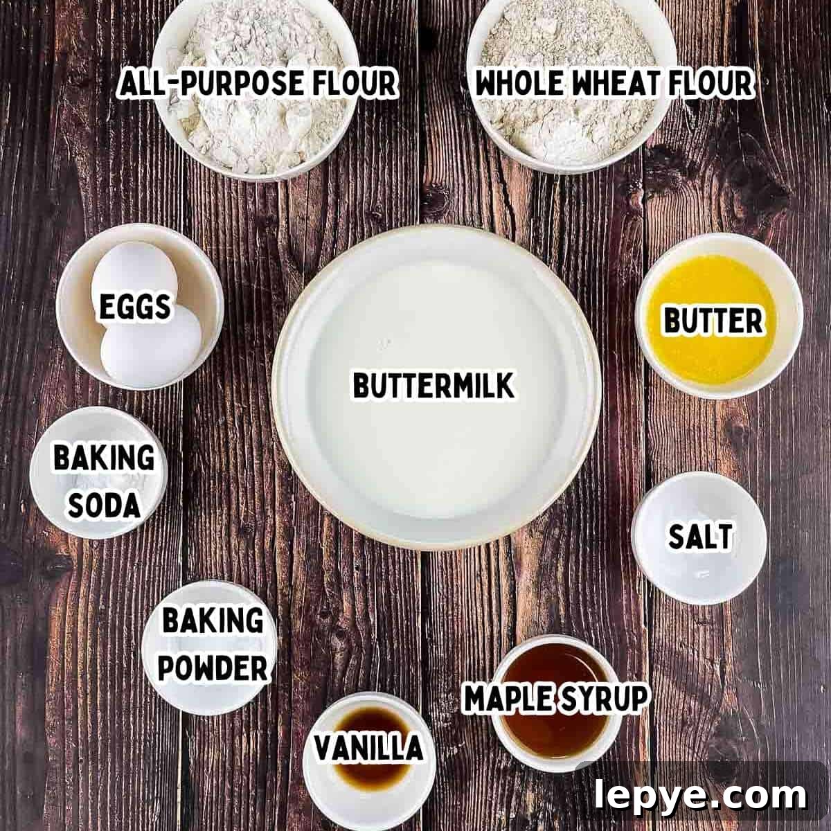 The ingredients for crispy Belgian waffles on a wooden table: Flour, eggs, baking soda and baking powder, buttermilk, butter, salt, vanilla, and maple syrup.