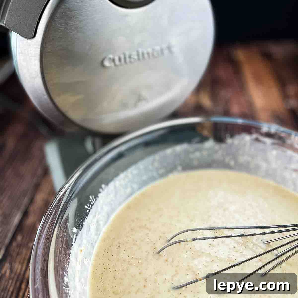 The mixed waffle batter in a bowl with a whisk and the Belgian waffle maker in the background.