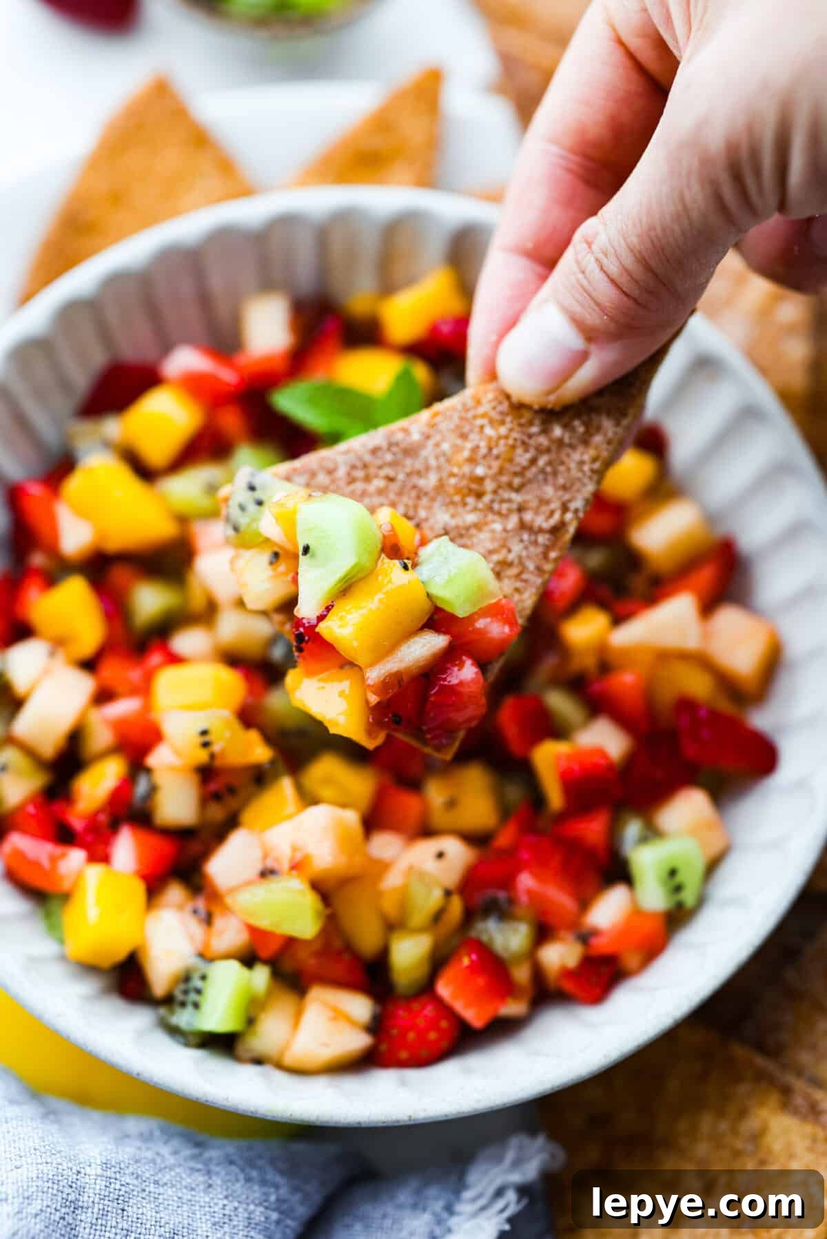 Vibrant fruit salsa is a fresh, juicy twist on your favorite snack! Sweet berries, tropical mango, and a splash of lime always steal the spotlight! Especially when served with crispy cinnamon chips! Someone taking a scoop of fruit salsa with a cinnamon sugar chip.