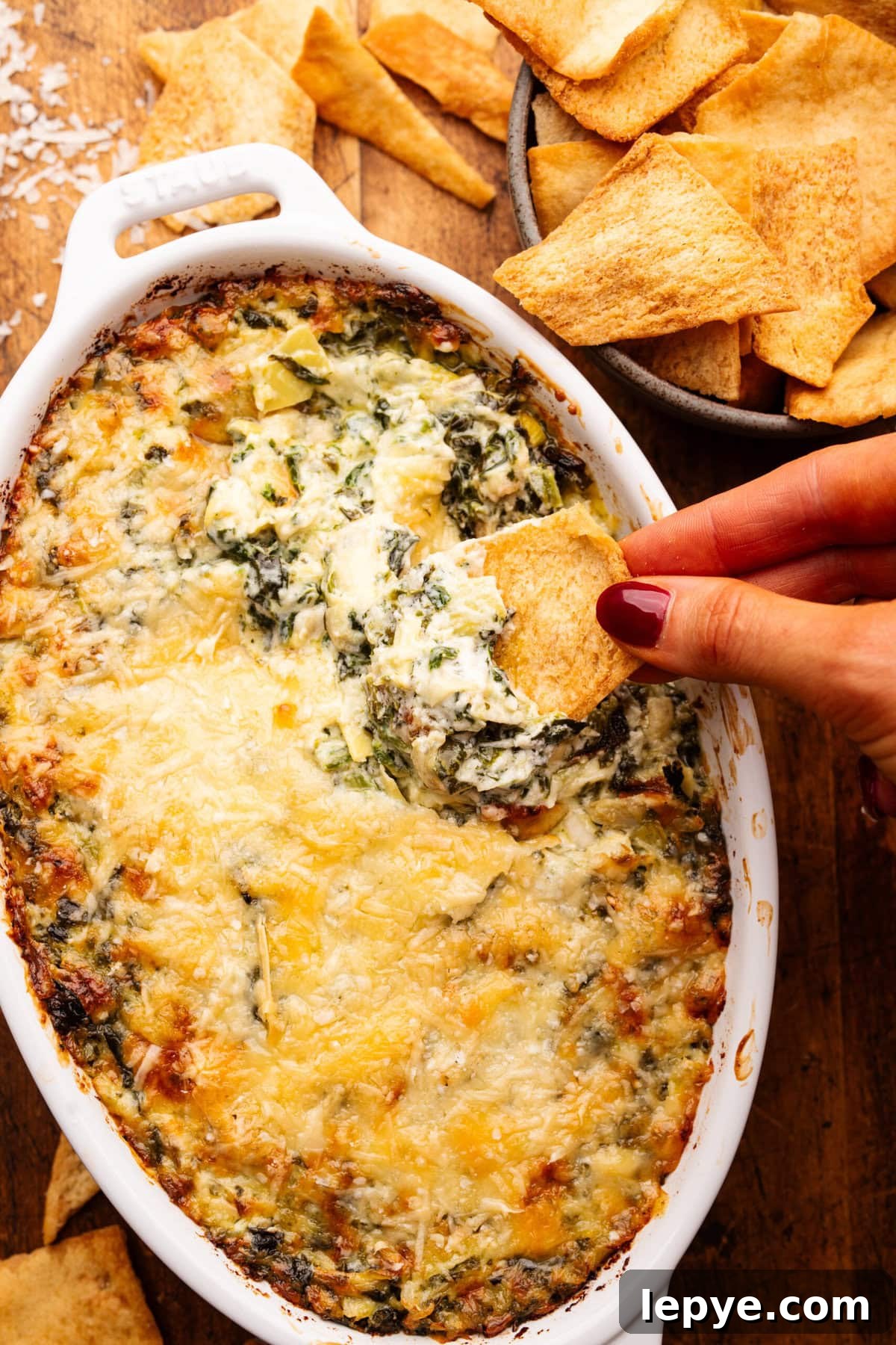 Creamy Baked Spinach and Artichoke Dip Recipe 2 A close up of a hand dipping a pita chip into a white oval dish with baked artichoke dip in it. Around the dip is a bowl of pita chips.
