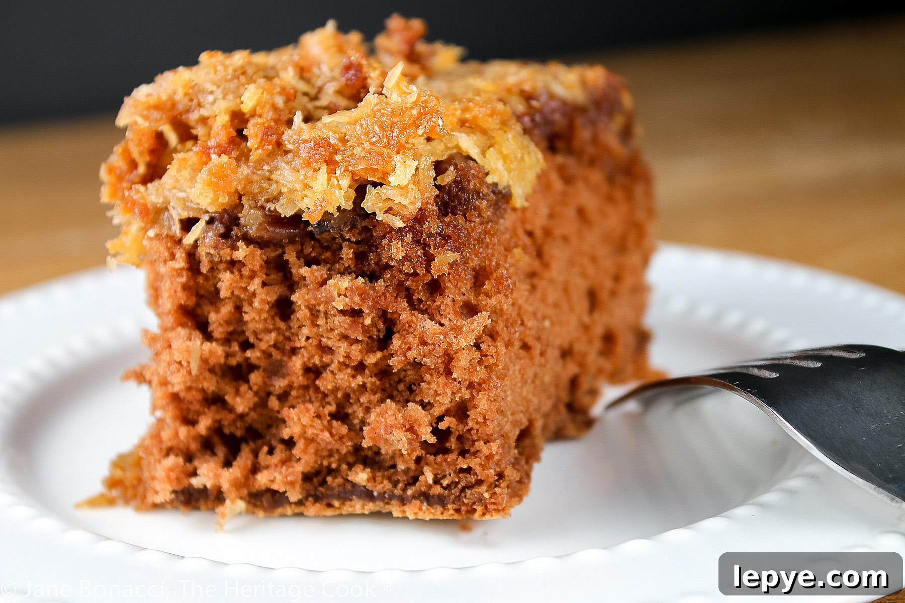 Square piece of cake with coconut/pecan frosting on a white plate; Cockeyed German Chocolate Cake © 2023 Jane Bonacci, The Heritage Cook.