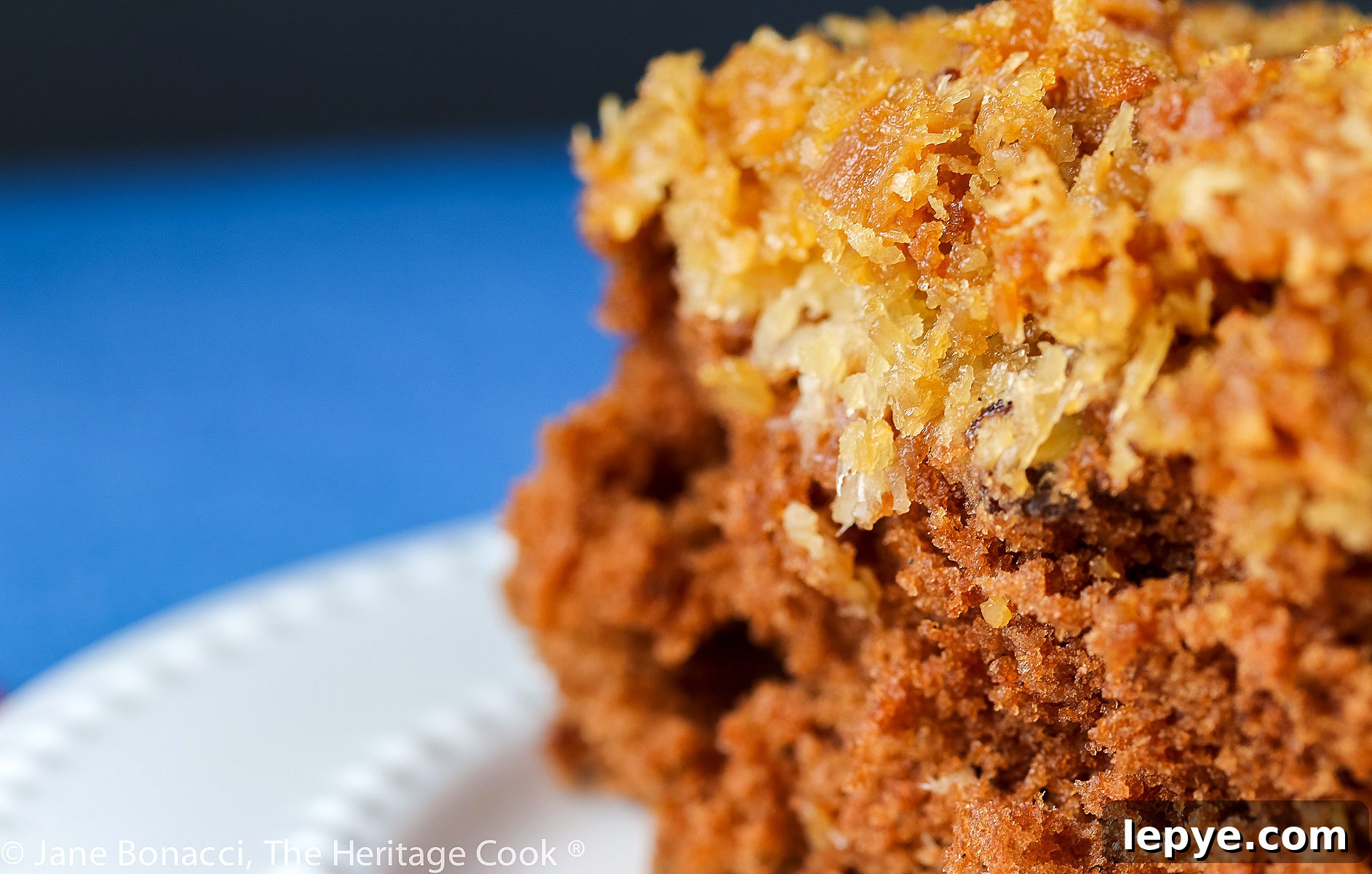 Square piece of cake with coconut/pecan frosting on a white plate; Cockeyed German Chocolate Cake © 2023 Jane Bonacci, The Heritage Cook.