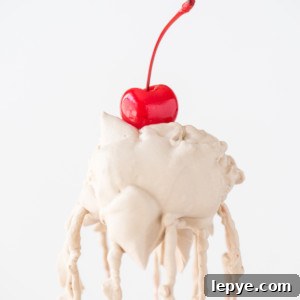 A bright red cherry sits atop a scoop of melting vanilla ice cream, with root beer whipped cream swirled on top and drips running down the sides against a plain white background.