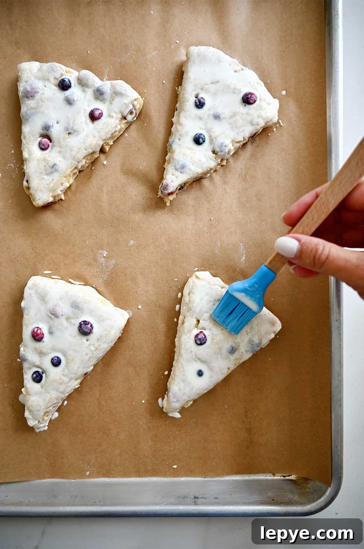 Using a pastry brush to brush heavy cream atop unbaked cranberry scones on a parchment paper-lined baking sheet.