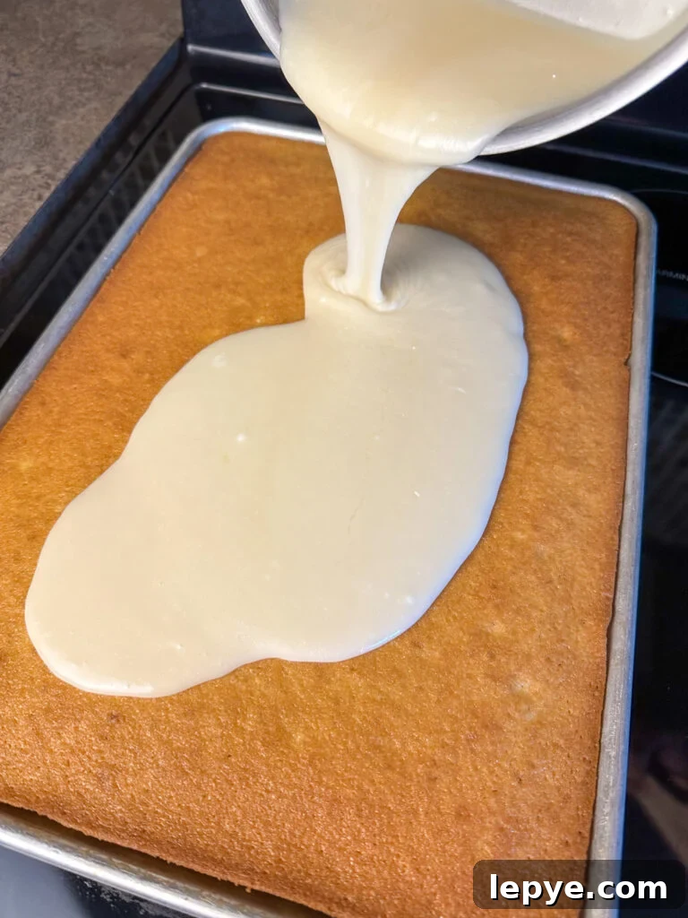 Southern-Style White Texas Sheet Cake Recipe 10 White icing being poured out of a sauce pan on top of a warm white Texas sheet cake.