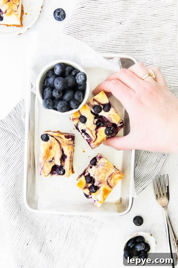 overhead shot of a hand picking up 1 of 3 blueberry cheesecake bars on a parchment paper lined 1/8 sheet pan