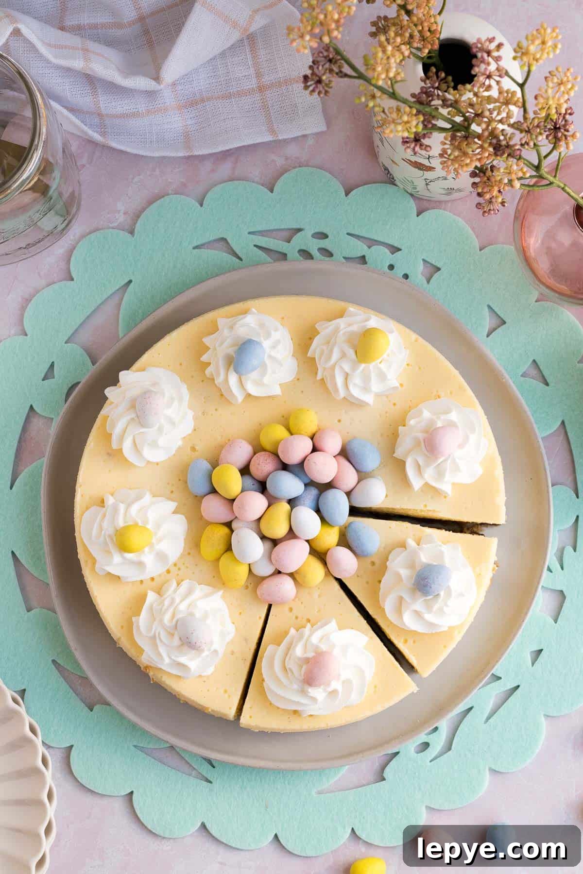 Top view of an Easter egg cheesecake with whipped cream and mini eggs piled on top and 2 slices cut sitting on a green Easter placemat