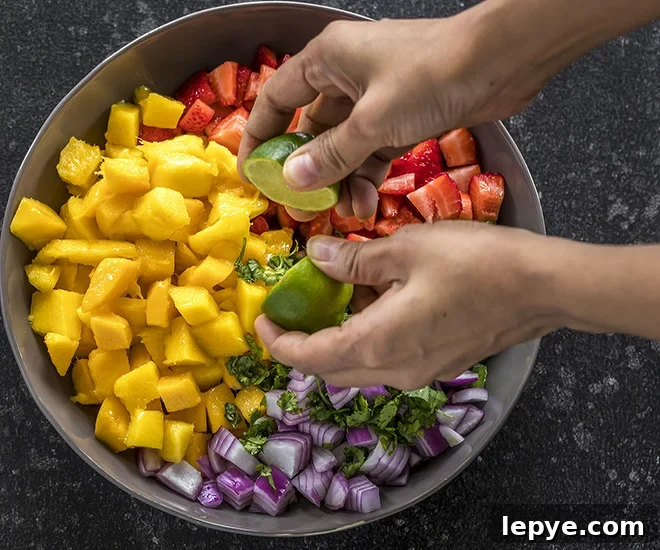 a pair of hand squeezing lime juice into a bowl of strawberry mango salsa