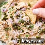 Dairy free smoked salmon dip in a small shallow ceramic speckled bowl, garnished with capers, fresh dill, everything but the bagel seasoning, and square gluten free crackers tucked into the dip and a hand dipping a cracker into the dip to lift it out.