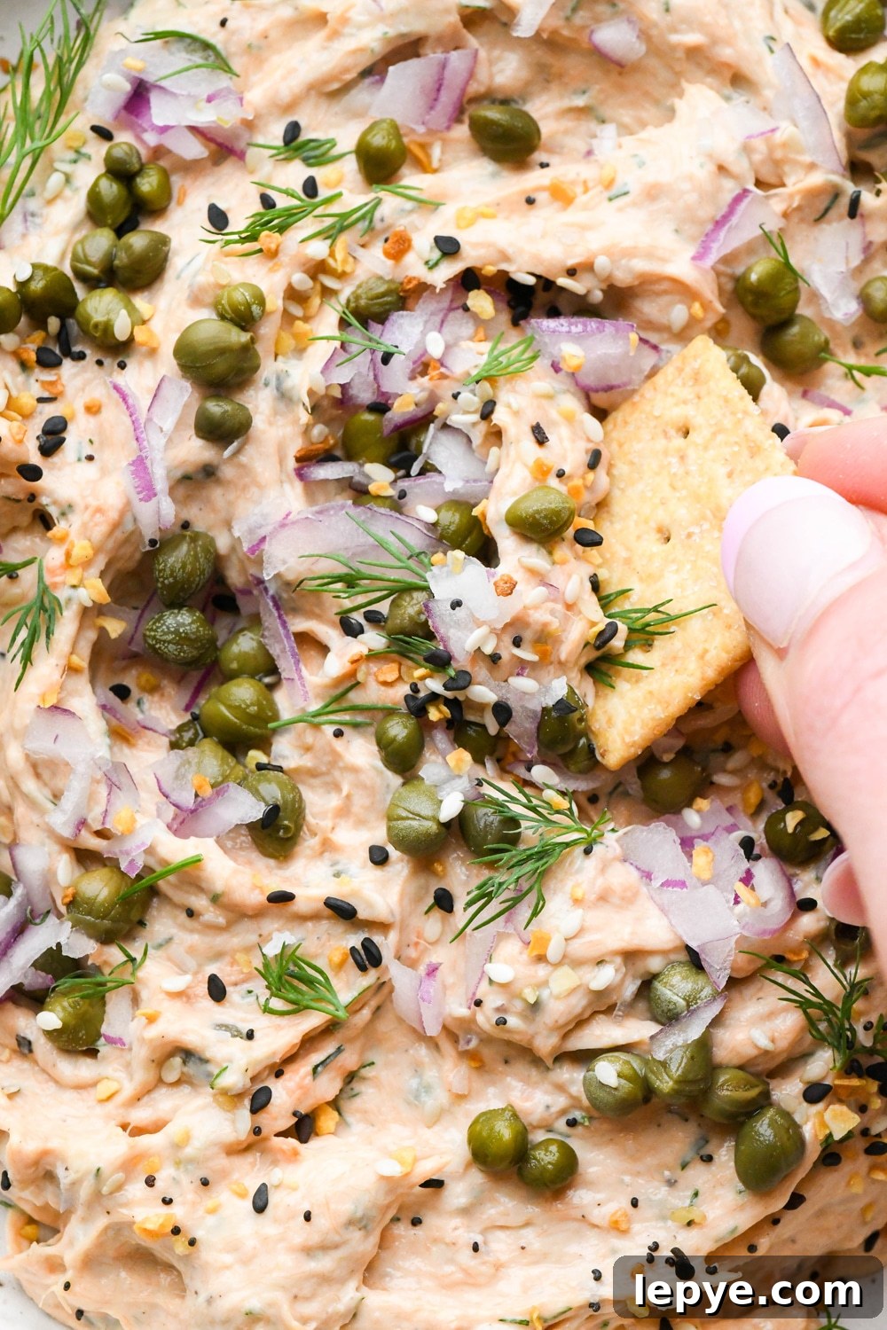Dairy free smokes salmon dip in a small shallow ceramic speckled bowl, garnished with capers, fresh dill, everything but the bagel seasoning, and square gluten free crackers tucked into the dip and a hand dipping a cracker into the dip to lift it out.