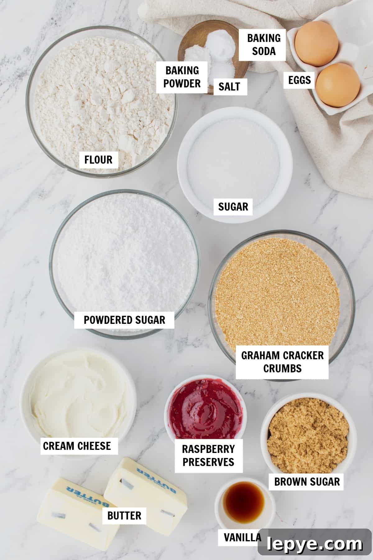 Raspberry Cheesecake Cookie Recipe — Soft, Jam-Filled Treats 3 All of the ingredients you need for Raspberry Cheesecake Cookies sitting on a white countertop including butter, sugar, eggs, vanilla, flour, graham cracker crumbs, salt, baking powder, baking soda, cream cheese and raspberry preserves.