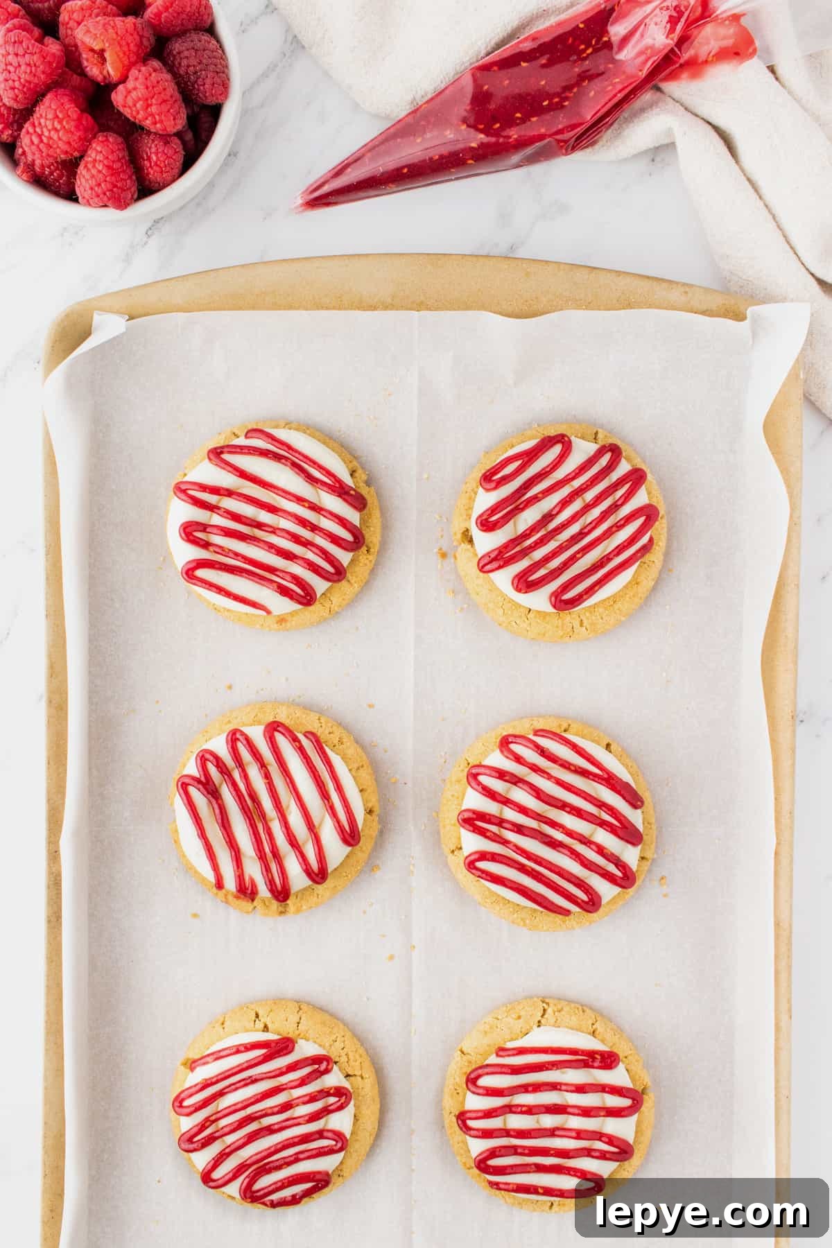 Raspberry Cheesecake Cookie Recipe — Soft, Jam-Filled Treats 5 Raspberry cheesecake cookies on a baking sheet after the raspberry preserves are drizzled on top of the cream cheese frosting.