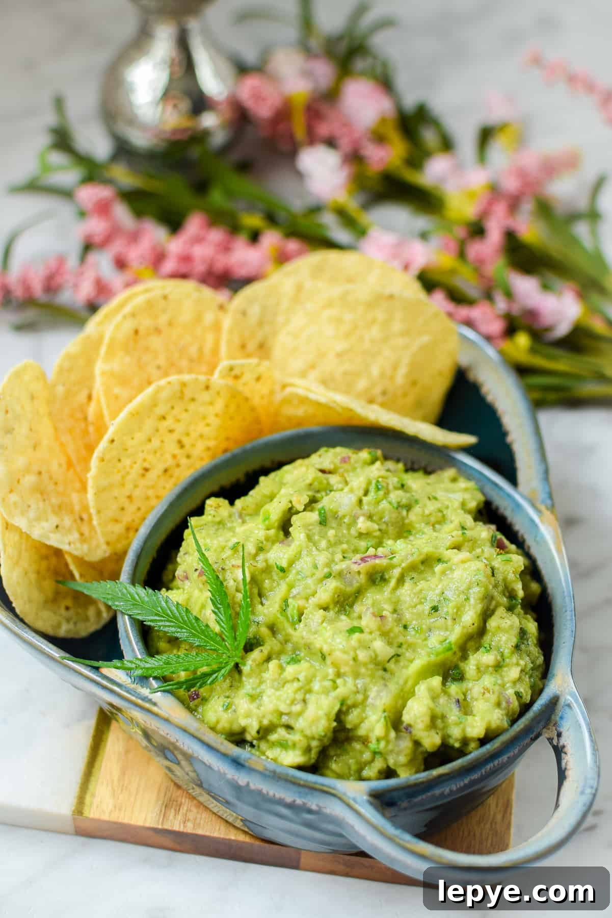 Cannabis-Infused Guacamole Recipe: Fast Homemade Pot Guac 2 A blue bowl filled with guacamole garnished with a cannabis leaf paired with round tortilla chips
