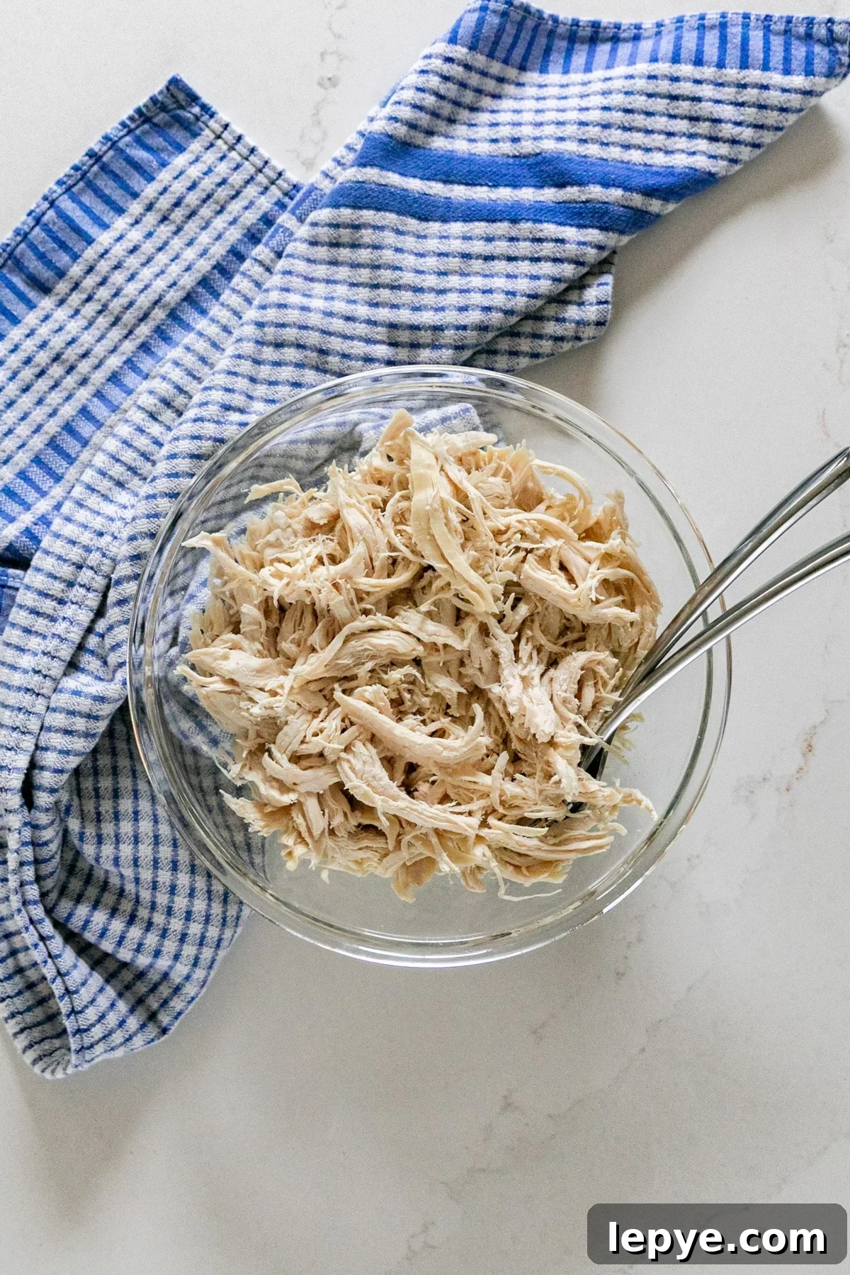 Shredded Chicken Recipes: Crockpot and Instant Pot Guide 2 An overhead image of shredded chicken breast in a glass bowl with a blue checked towel next to it and a couple of forks in it.