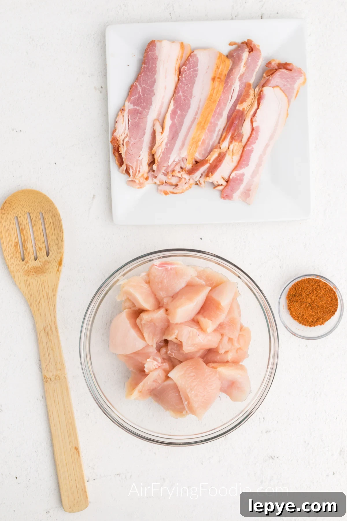 Crispy Bacon-Wrapped Chicken Bites Made in the Air Fryer 4 Bacon on a white plate, seasonings in a small container, and chicken pieces in a bowl.