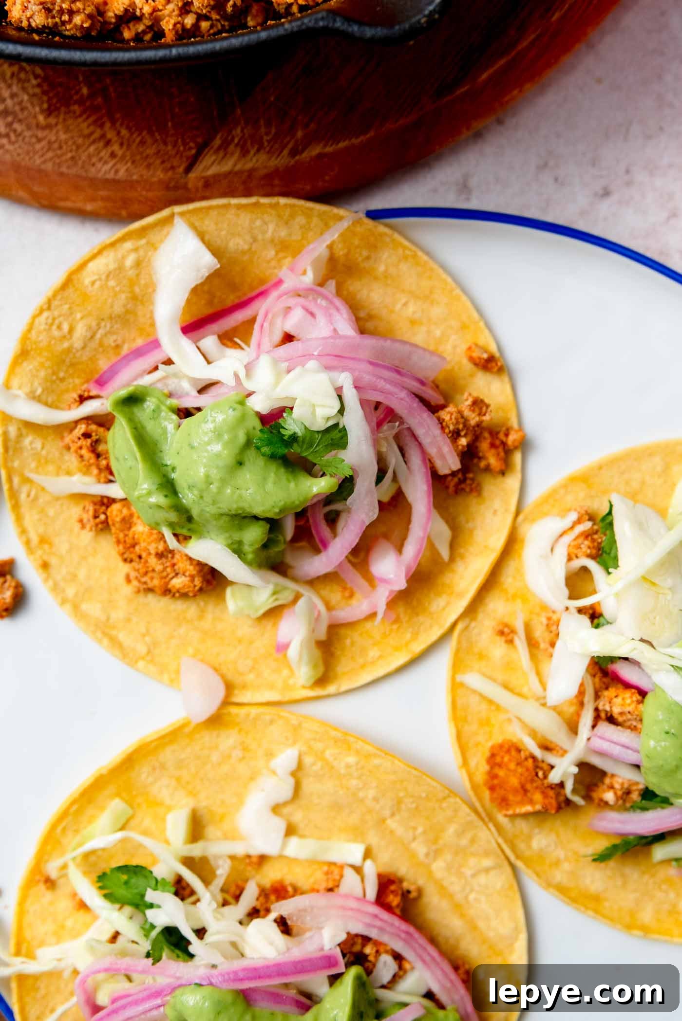 Three tofu tacos topped with cabbage, avocado sauce and pickled onion on a plate.