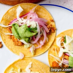 Three tofu tacos topped with cabbage, avocado sauce and pickled onion on a plate.