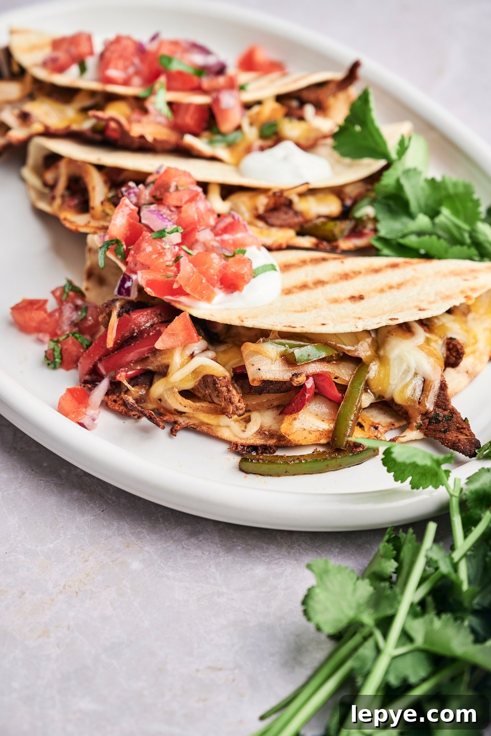 Three steak quesadillas are on a white serving platter. The quesadillas are topped with pico de gallo and sour cream. Fresh cilantro is on the platter as well.