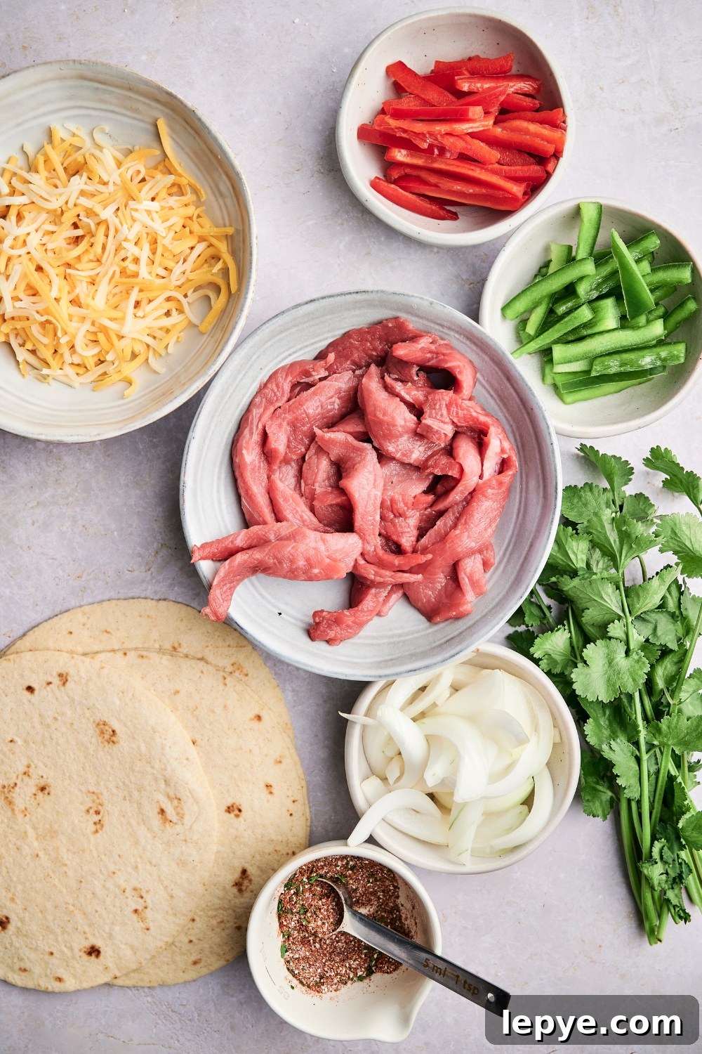 Steak strips, bell peppers, shredded cheese, tortillas, onions, taco seasoning, and fresh cilantro are in separate containers on a counter top.