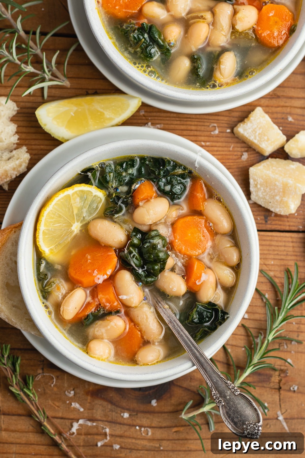 This is a picture of the lemony white bean soup in a bowl with a spoon, parmesan, bread and a lemon wedge.