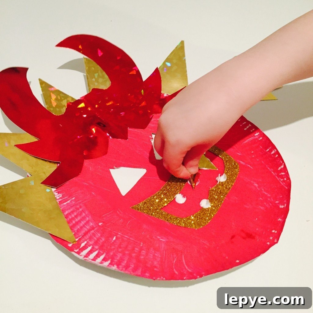 Chinese Dragon Mask Craft for Kids: Step-by-Step Paper Mask Tutorial 9 child sticking decorations onto a dragon mask craft