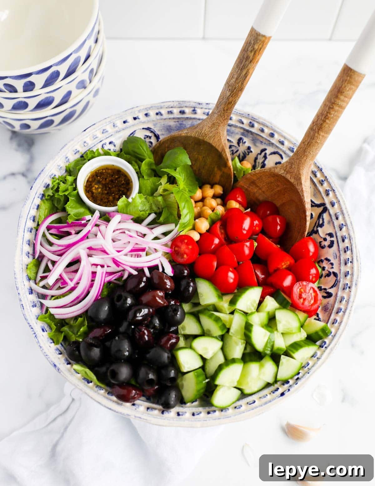 A salad with fresh vegetables, olives, lettuce, chickpeas, and a vinaigrette in a large blue and white bowl with salad tongs sticking out of it