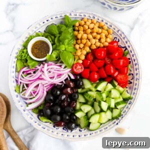 A large salad with lettuce, basil, chickpeas, tomatoes, cucumbers, olives, red onion, and a small white dish filled with dressing