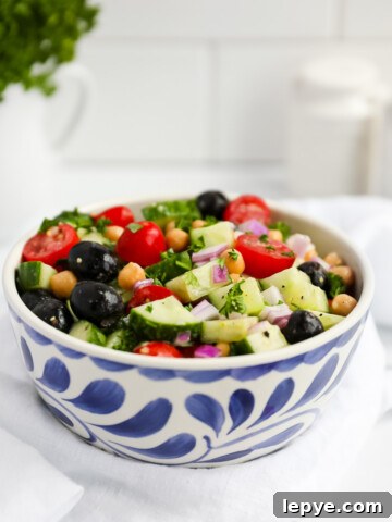 Vegan Mediterranean cucumber salad in a blue and white bowl including onion, tomatoes, olives, cucumber, chickpeas, and herbs