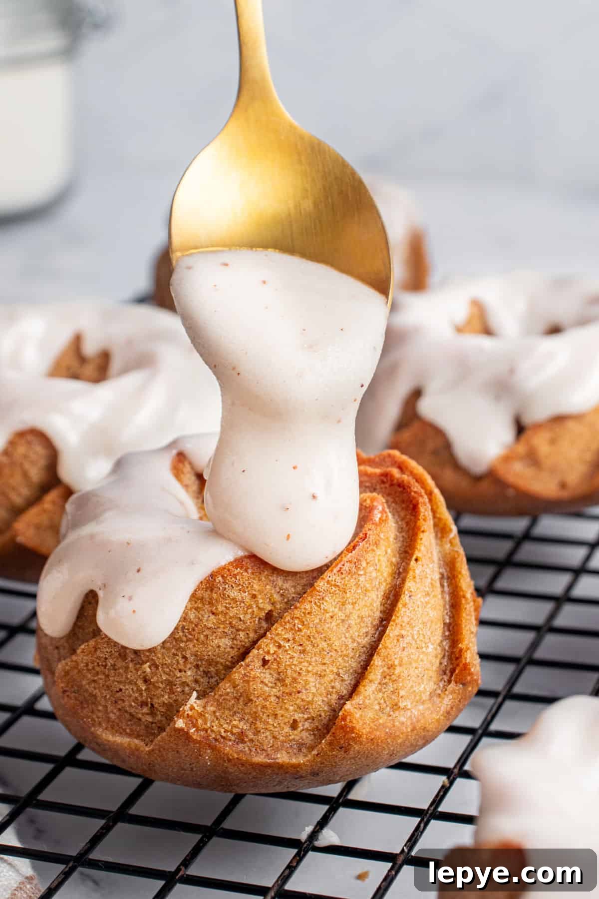 Glazing mini bundt pumpkin cake on a black wire rack. Golden spoon, gray background.