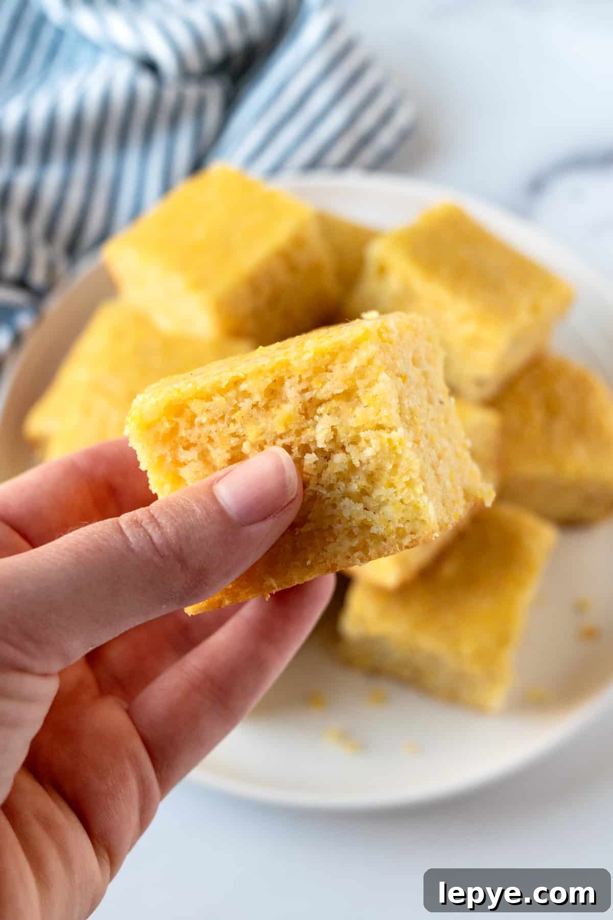 Honey-Glazed Cornbread Recipe with Golden Crust and Sweet Drizzle 7 A hand holding a piece of cornbread, with a plate of cornbread in the background.