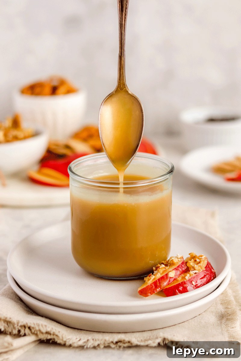 DIY Smooth Caramel Dip Recipe Without Condensed Milk 2 caramel sauce in a jar on a plate with apple slices on it. There is a spoon drizzling the caramel into the jar.