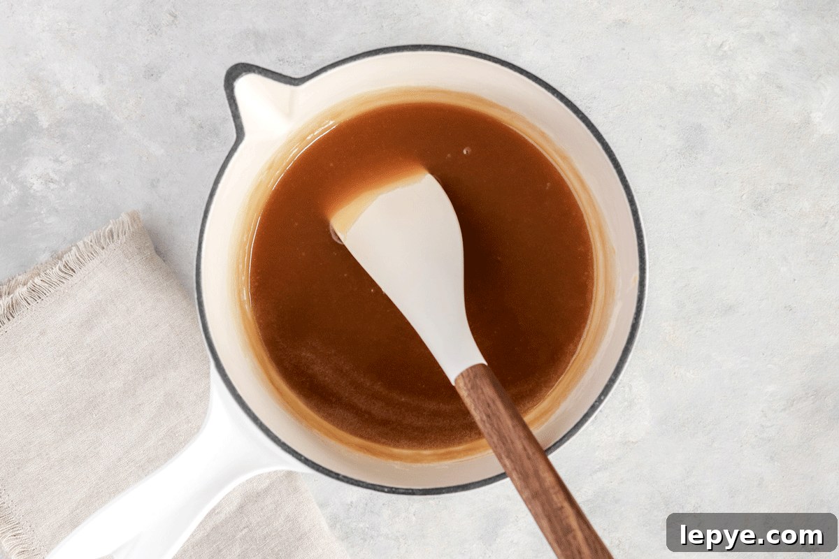 DIY Smooth Caramel Dip Recipe Without Condensed Milk 4 caramel sauce in a jar on a plate with apple slices on it. There is a spoon drizzling the caramel into the jar.