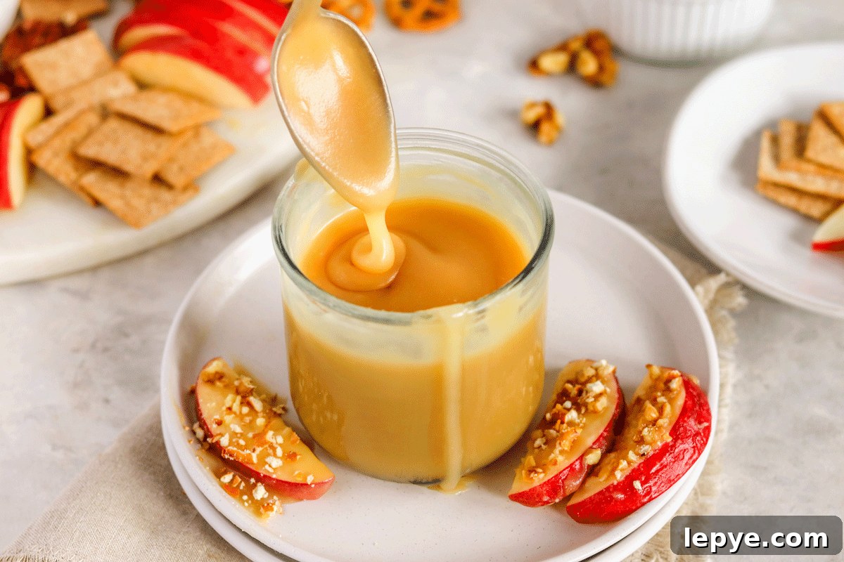 DIY Smooth Caramel Dip Recipe Without Condensed Milk 6 caramel sauce in a jar on a plate with apple slices on it. There is a spoon drizzling the caramel into the jar.
