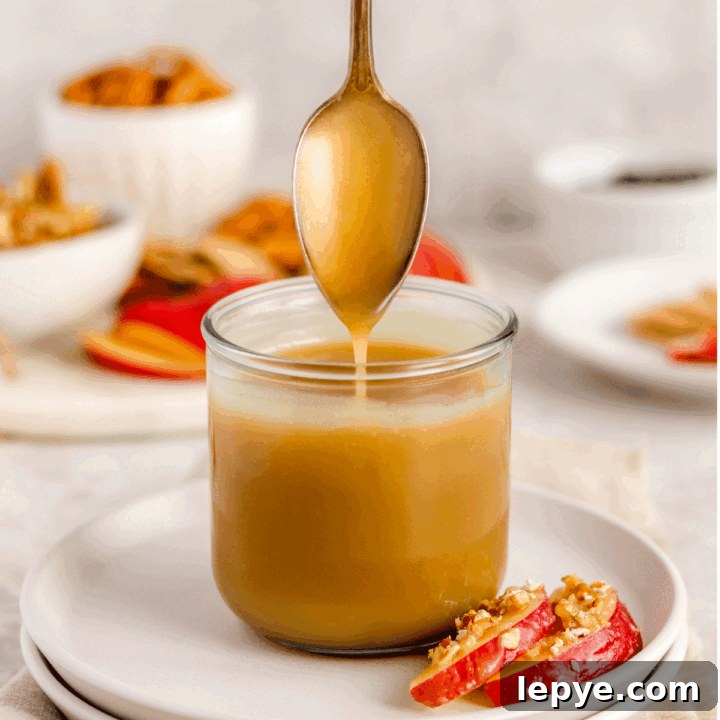 caramel sauce in a jar on a plate with apple slices on it. There is a spoon drizzling the caramel into the jar.